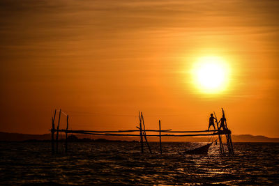Silhouette men on wooden structure fishing in sea against orange sky during sunset