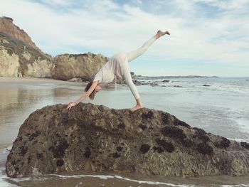 Woman doing yoga on rock at beach against sky