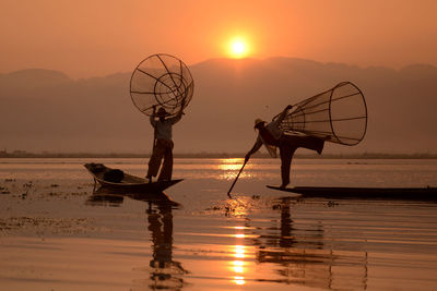 Fishermen fishing in lake against sky during sunset