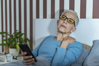Senior woman with blue-light blocking glasses lying in bed before sleeping, using smartphone.
