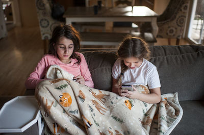 Friends sit on couch under blanket looking at their phones