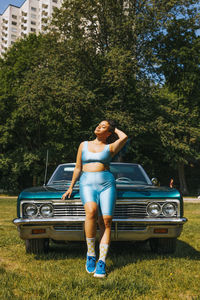 Young vuluptuous woman with hand in hair standing in front of car at sunny day