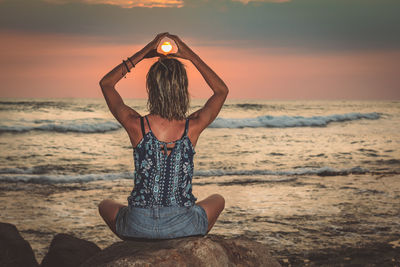 Woman doing yoga on rock at beach against sky during sunset