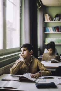 Thoughtful boy with hand on chin looking outside window while sitting in classroom