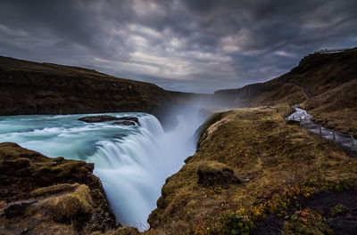 Idyllic view of waterfall by mountains against cloudy sky