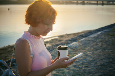 Mid adult woman using mobile phone while standing by river against sky during sunset