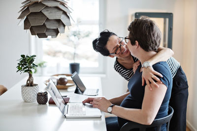 Smiling woman embracing girlfriend using laptop at table