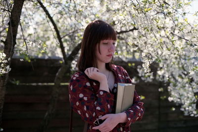 Woman holding book while standing by tree