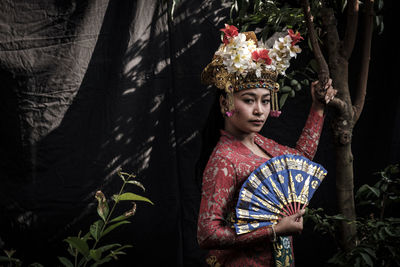 Portrait of beautiful young woman in traditional clothing holding hand fan while standing against trees