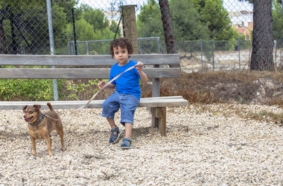 Four year old boy dressed in blue, in park with his little brown dog sitting on wooden bench