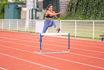 Full length of confident young hispanic sportswoman jumping over barrier while running on red track of sports stadium
