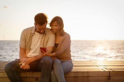 Smiling woman showing mobile phone to boyfriend while sitting on seat against sea during sunset