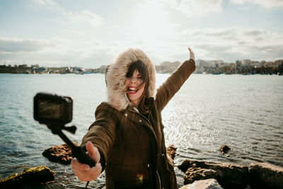 Happy woman taking selfie with camera while standing against lake