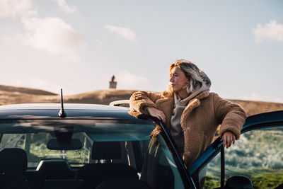 Young woman looking away while standing by car against sky