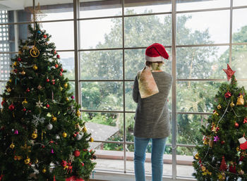 Back of standing caucasian senior woman alone at home looking outside window on christmas new year 