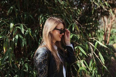 Smiling young woman wearing sunglasses while standing against plants in park