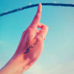 Cropped hand with deer tattoo touching rope against blue sky