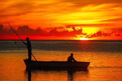 Silhouette men fishing in sea against orange sky