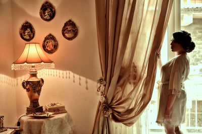 Side view of young woman standing by window in illuminated home