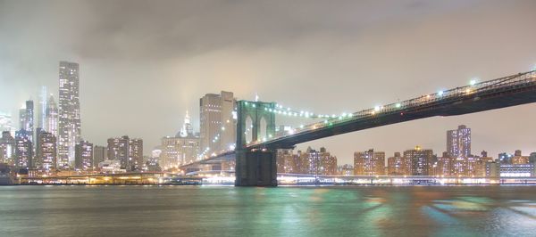 Illuminated bridge over river by buildings against sky at night