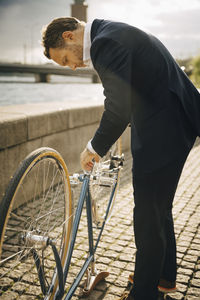 Side view of businessman repairing bicycle on footpath in city during sunny day