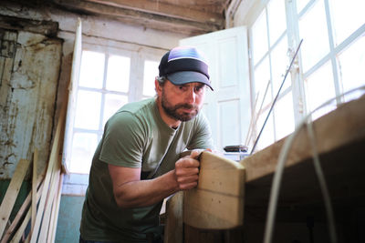 Concentrated bearded male worker in cap and t shirt making longboard at workbench in daylight