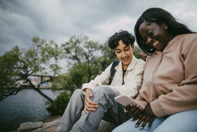 Low angle view of smiling teenage girl using smart phone while sitting with male friend