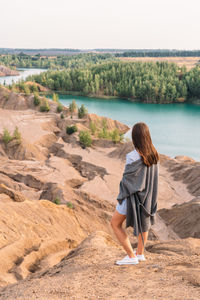 Beautiful unrecognizable girl in blue dress and grey cape stands with her back. sandy rocks