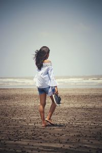 Rear view full length of woman holding shoes at beach against sky