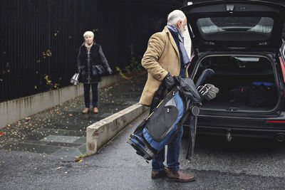 Senior man holding golf bag by car trunk while female partner standing on sidewalk during winter