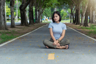 Portrait of young woman sitting on sidewalk against trees