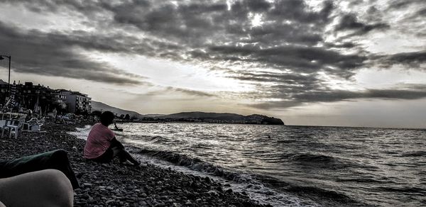 Woman sitting on rock at beach against sky during sunset