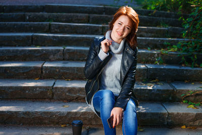  red-haired girl, dressed in a leather jacket with jeans, sits on the steps with a glass of coffee.