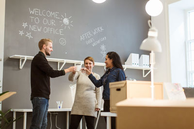 Low angle view of happy multi-ethnic colleagues toasting drinks while standing against wall at creative office