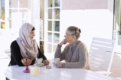 Female home caregiver and senior woman conversing while having coffee on porch