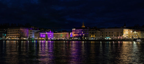 Illuminated buildings by river against sky at night