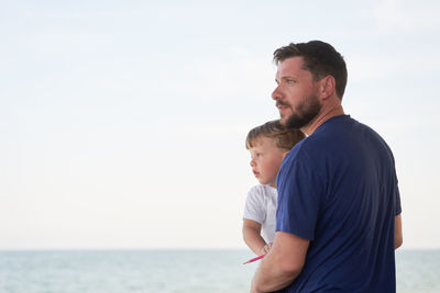 Father and son looking away at shore against sky
