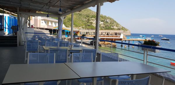 Empty chairs by swimming pool in sea against sky