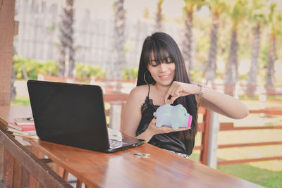 Young woman putting coin in piggy bank while sitting at cafe