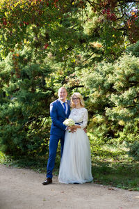 Portrait of smiling bride and groom standing amidst plants on roadside at park