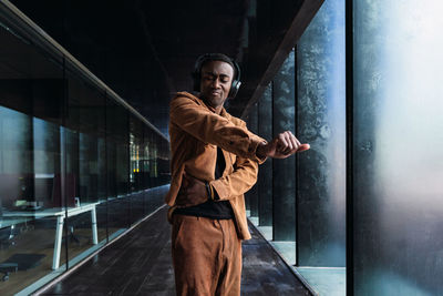 Stylish african american male dancing and listening to music in wireless headphones while standing near modern building with glass wall