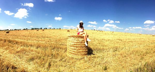 Full length of young woman sitting on hay bale against sky during sunny day