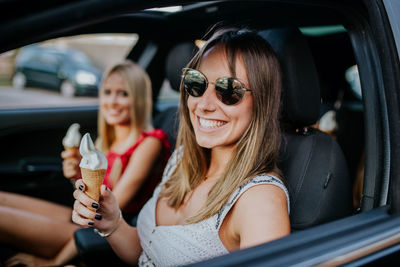 Cheerful millennial female friends eating ice cream cone in car during road trip in summer day