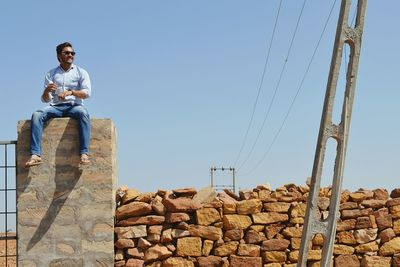 Low angle view of man sitting on retaining wall against sky