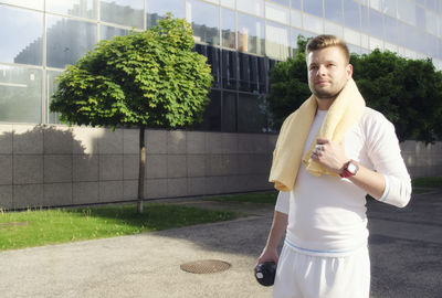 Young male jogger with towel around neck while standing on street by modern building in city