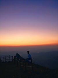 Man sitting on fence at observation point against sky during sunset