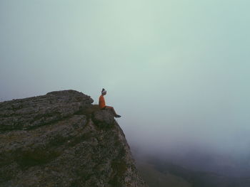 Side view of woman sitting on mountain cliff during foggy weather
