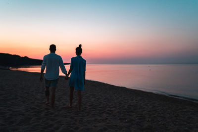 Rear view of friends standing on beach against sky during sunset