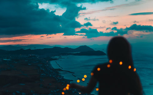 Rear view of silhouette woman with illuminated string light standing by sea against sky