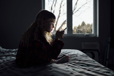 Side view of girl using mobile phone while sitting on bed at home